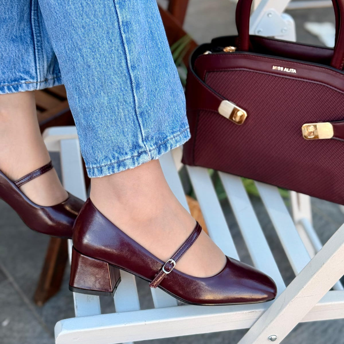 Person wearing maroon shoes and holding a matching handbag on a chair.