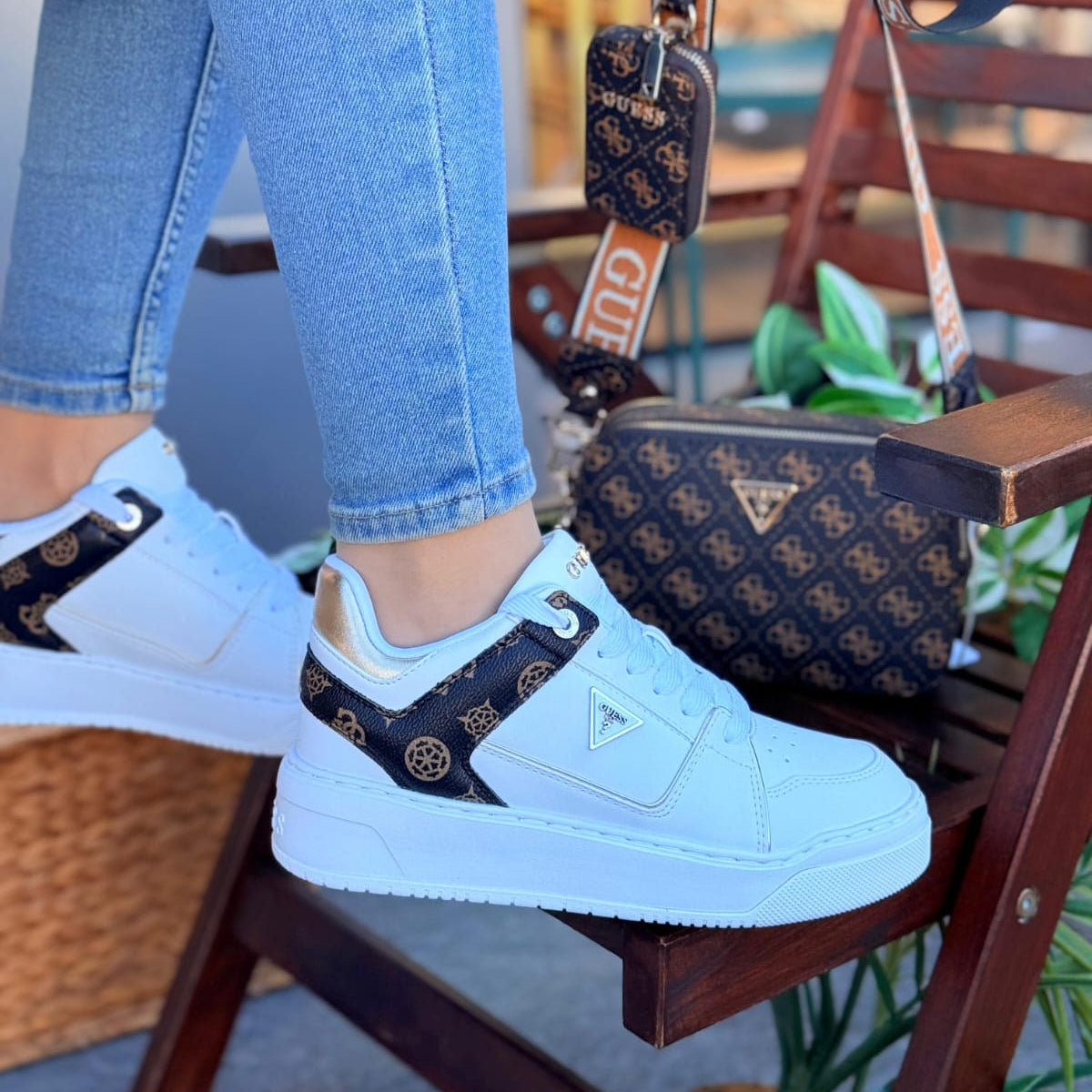 White sneakers with black pattern worn by a person sitting on a chair with a patterned bag in the background.