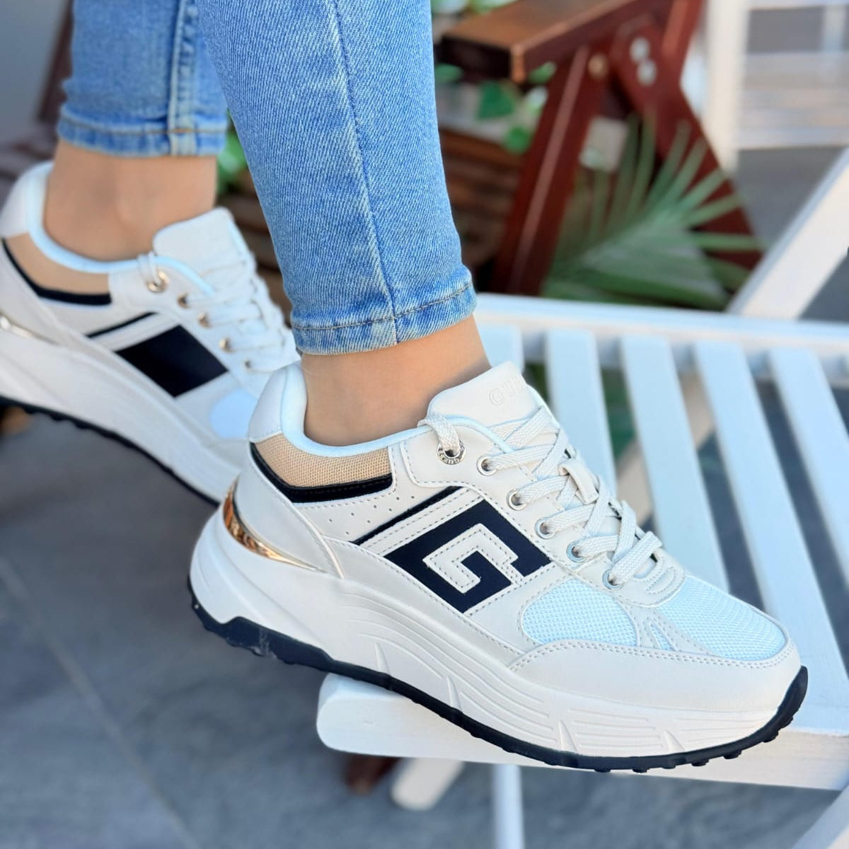 White sneakers with black and gold accents worn by a person sitting on a chair.