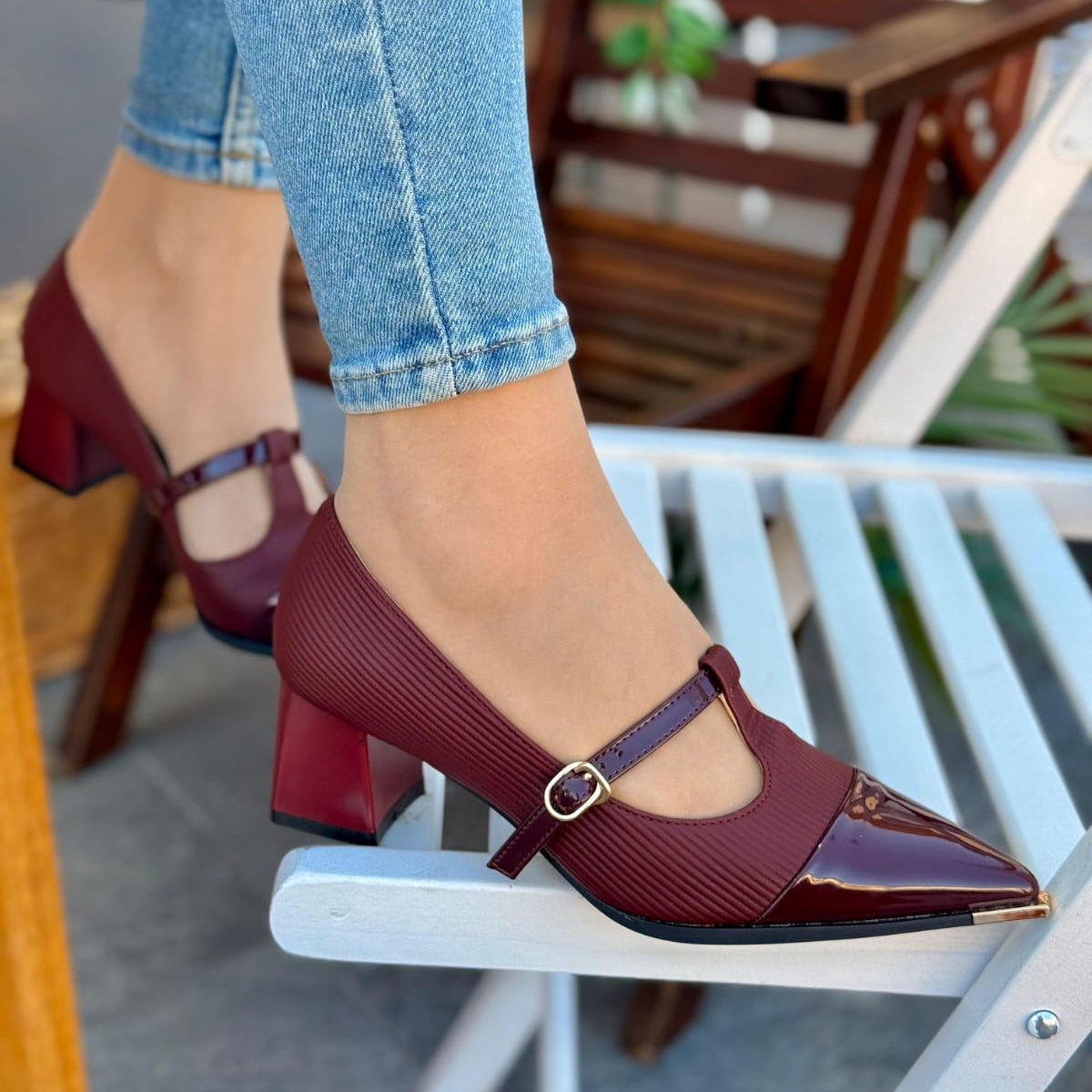 Maroon high-heeled shoes with a gold buckle worn with blue jeans on a chair.