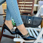 Person wearing black high-heeled shoes with studded details, sitting on a chair with a Guess handbag.