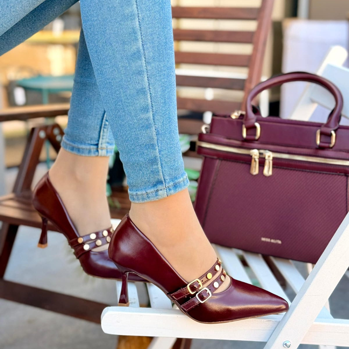 Maroon high-heeled shoes with gold buckles worn by a person sitting outdoors.