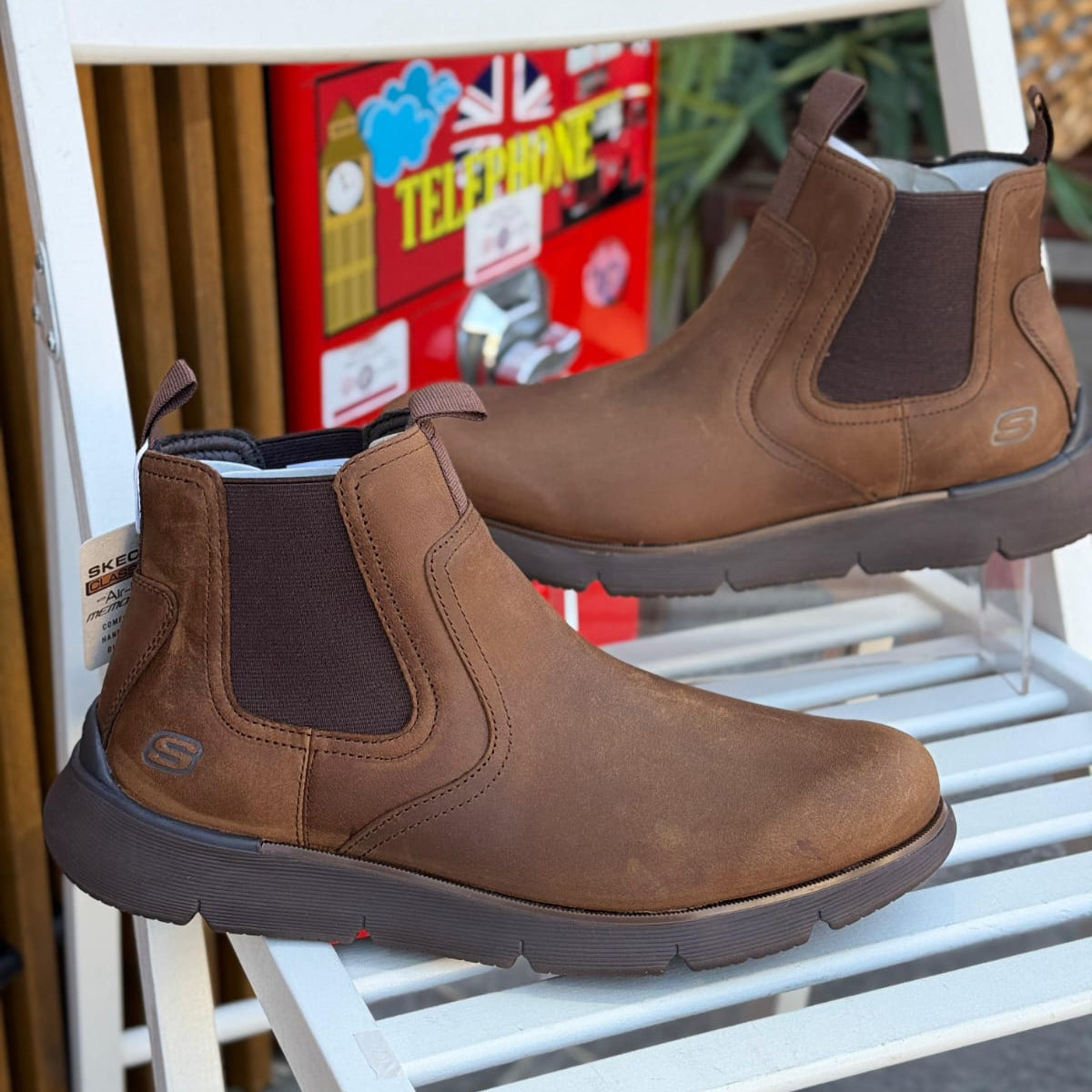 Pair of Skechers Augustino Paulo brown leather men’s boots displayed outdoors with plants in background.