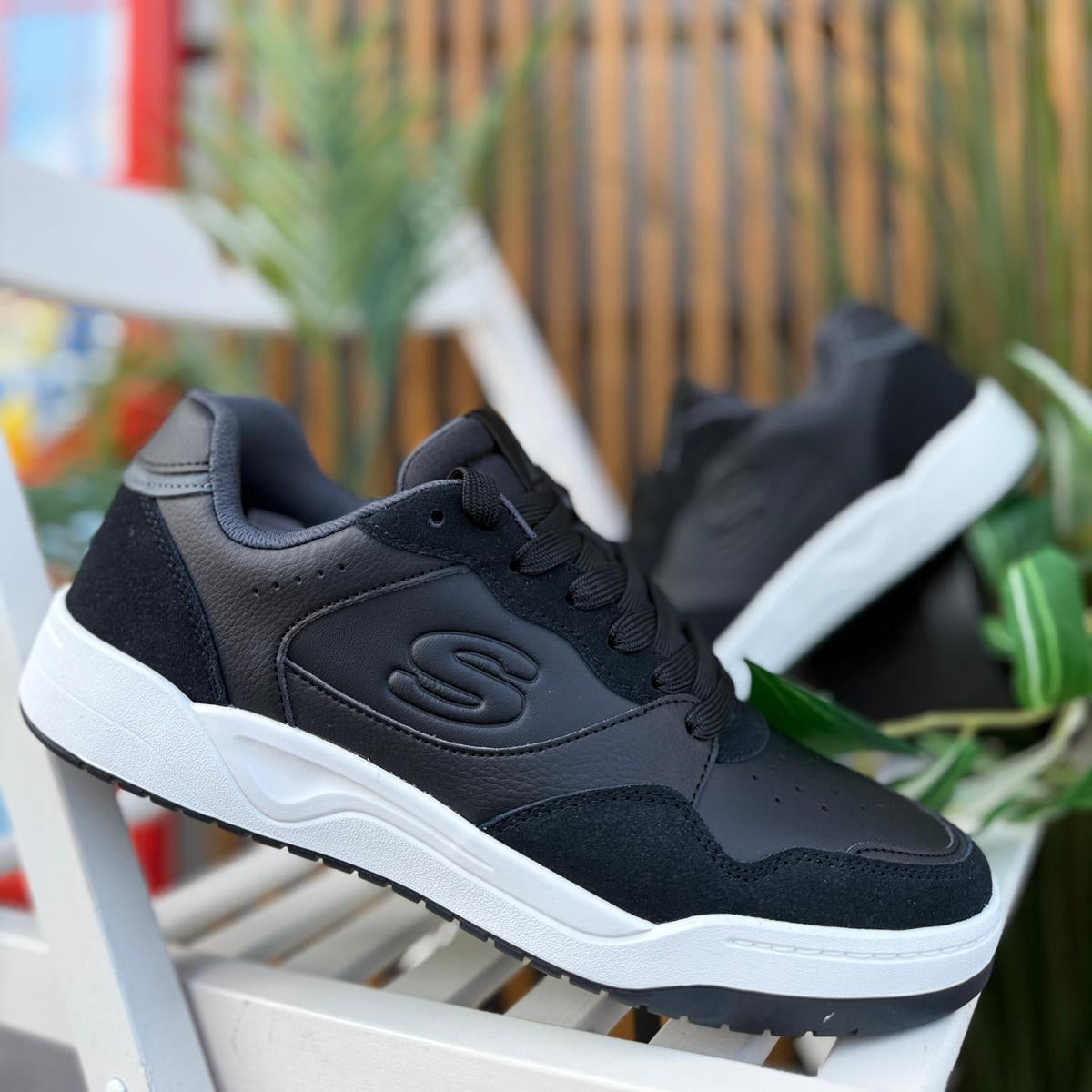 Pair of Skechers Koopa Volley Low Varsity black men’s sneakers displayed outdoors with plants in background.