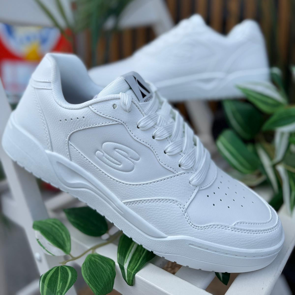 Pair of Skechers Koopa Volley Low Varsity men’s white sneakers displayed outdoors with plants in background.