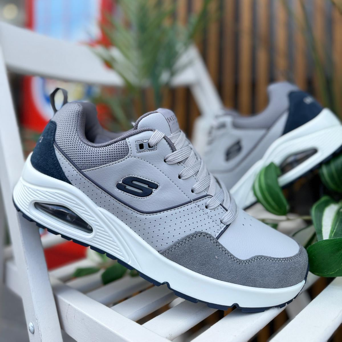 Pair of Skechers UNO Retro One men’s gray sneakers displayed outdoors with plants in background.