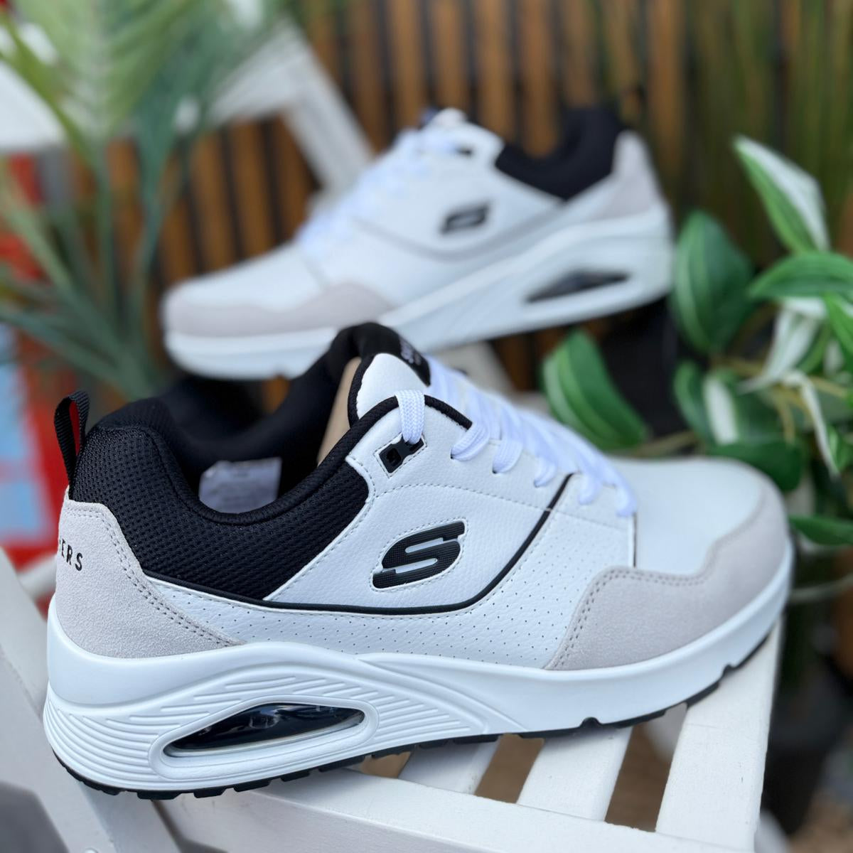 Pair of Skechers UNO Retro One men’s white-black sneakers displayed outdoors with plants in background.