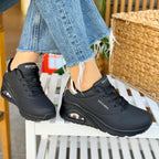 Woman wearing Skechers Uno Shimmer Away black sneakers with rose gold details sitting outdoors on a white chair.