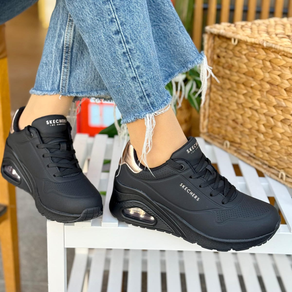 Woman wearing Skechers Uno Shimmer Away black sneakers with rose gold details sitting outdoors on a white chair.