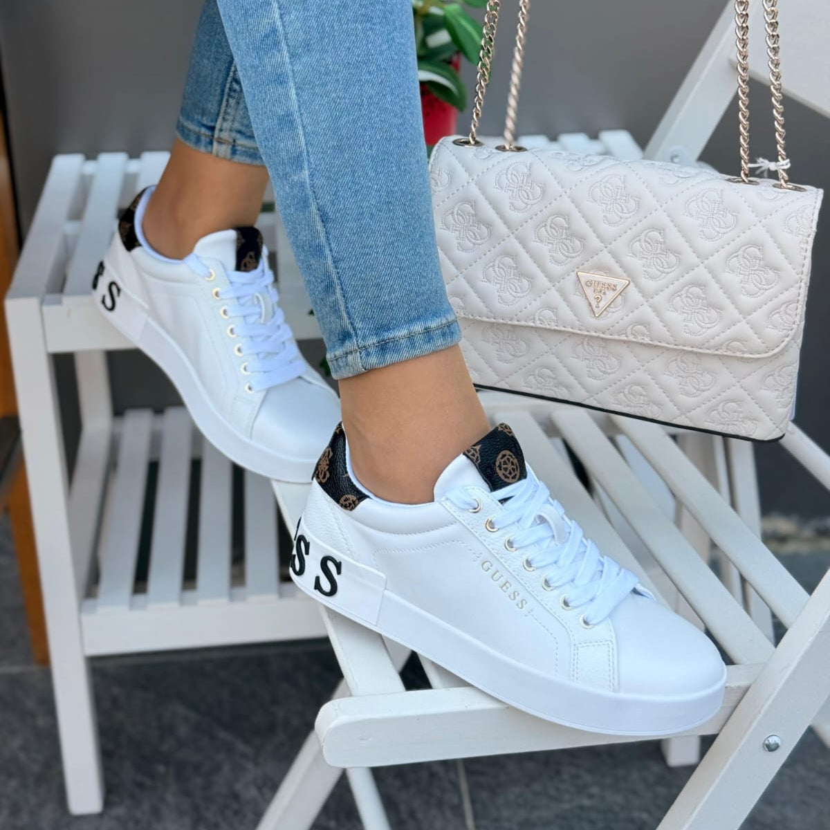 White sneakers with black accents worn by a person sitting on a white chair, with a light gray handbag in the background.