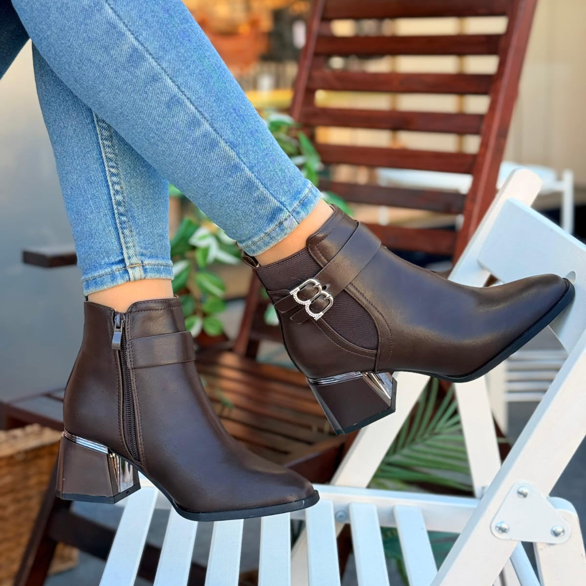 Brown ankle boots with a silver buckle worn by a person sitting on a white chair.