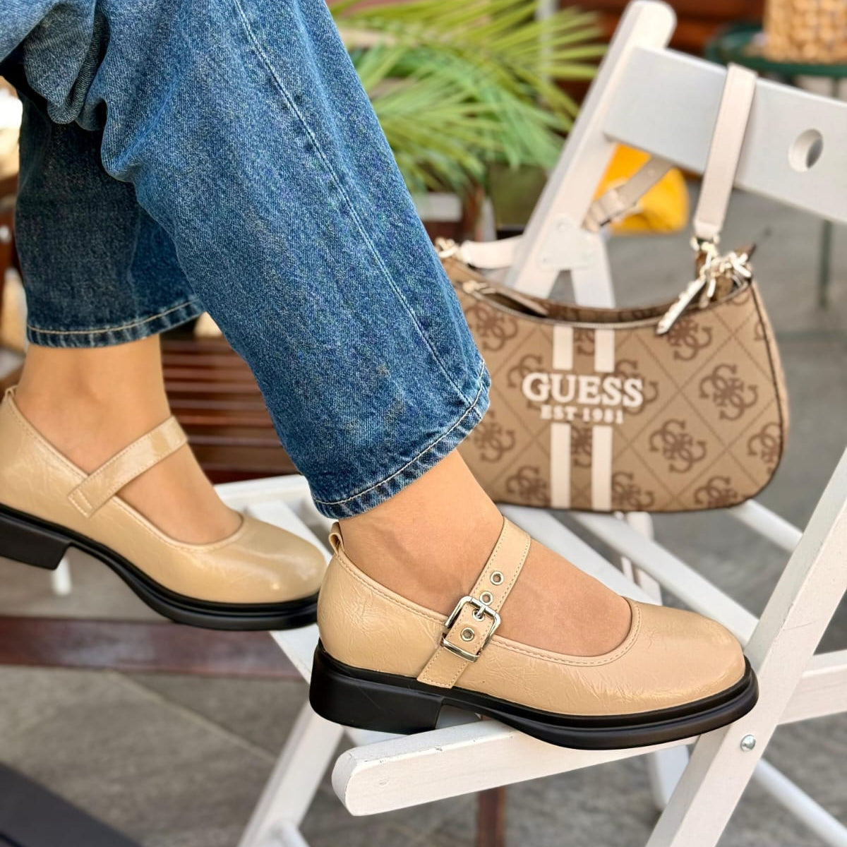 Beige shoes with black soles worn by a person sitting on a chair, with a Guess handbag in the background.