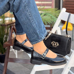 Person wearing black shoes with a gold buckle, sitting on a chair with a black handbag featuring a gold chain.