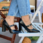 Person wearing black high-heeled shoes with buckles sitting on a chair.